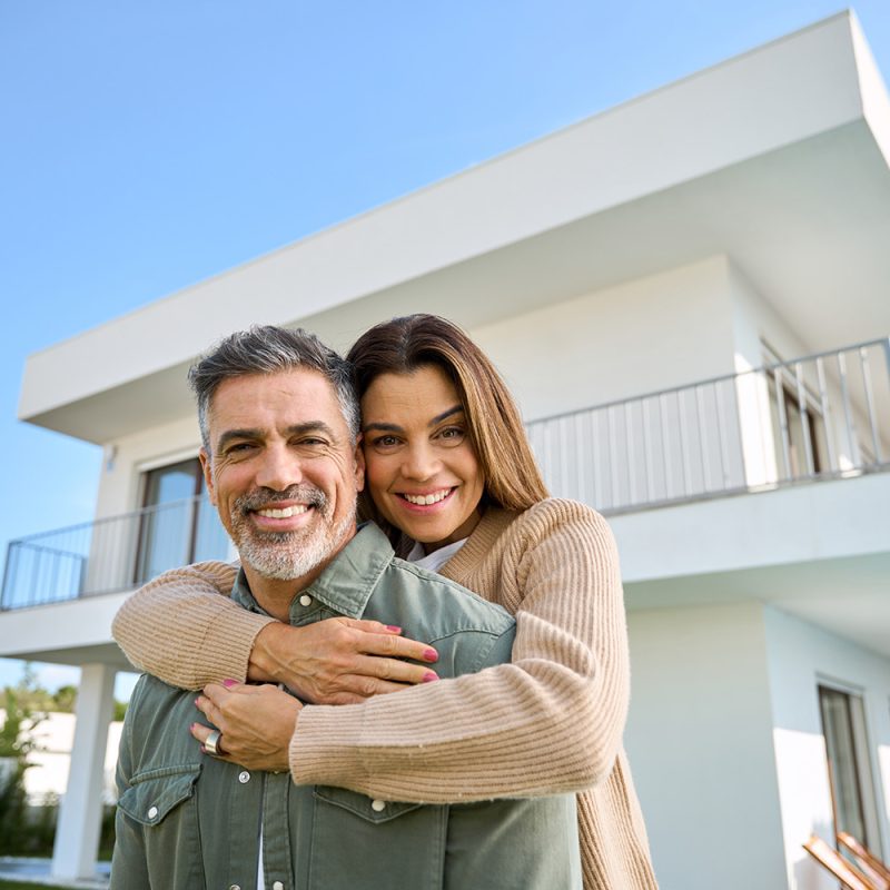 Happy smiling mature older family couple new property buyers modern dream luxury villa owners standing outside house, affectionate senior middle aged man and woman in love hugging outdoor. Portrait.